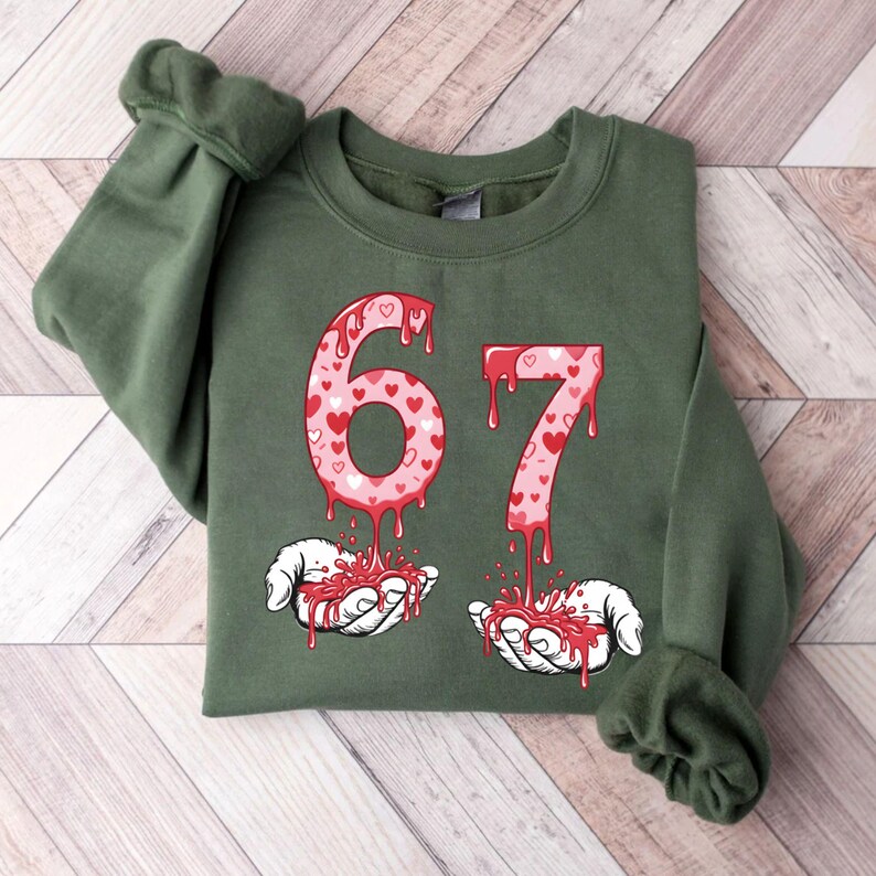 May include: An olive green sweatshirt featuring the number "67" in a dripping style. The numbers are filled with pink hearts. Below the numbers are two hands holding dripping red liquid.