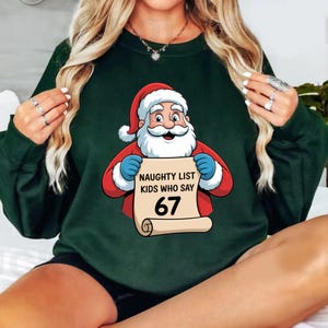 May include: Dark green sweatshirt featuring a Santa Claus graphic holding a scroll. The scroll reads "NAUGHTY LIST KIDS WHO SAY 67". Santa is wearing a red suit and hat with white trim and blue gloves. A festive holiday design.