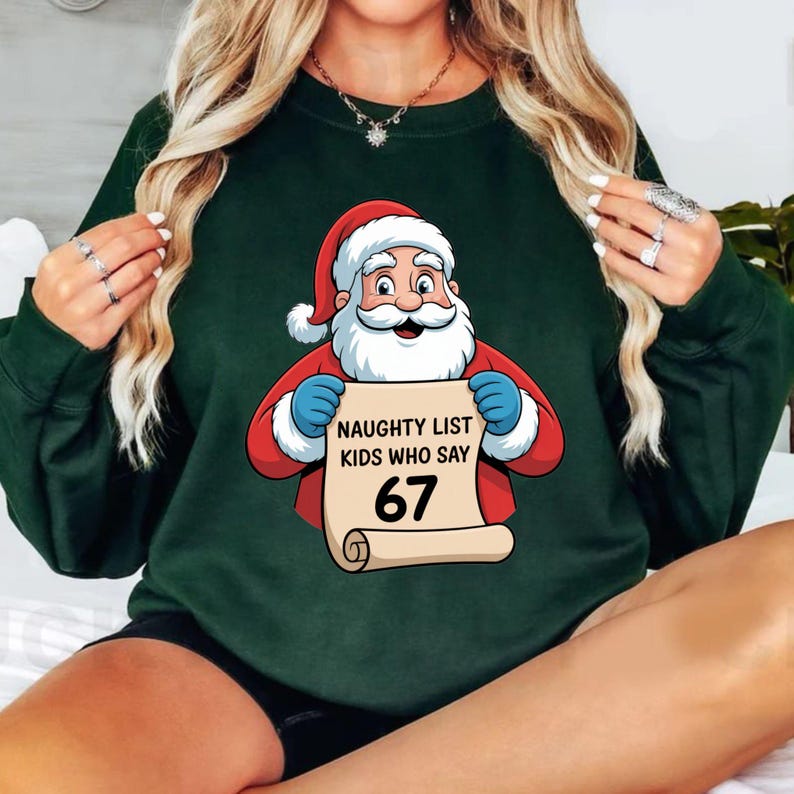 May include: Dark green sweatshirt featuring a Santa Claus graphic holding a scroll. The scroll reads "NAUGHTY LIST KIDS WHO SAY 67". Santa is wearing a red suit and hat with white trim and blue gloves. A festive holiday design.