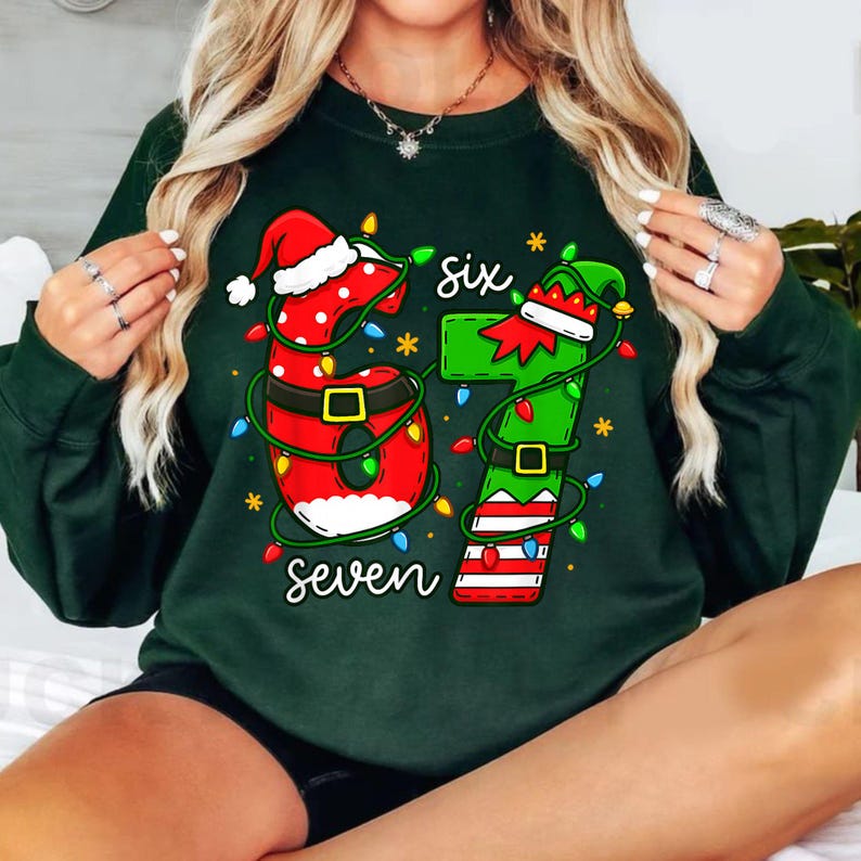 May include: Dark green sweatshirt with a festive Christmas design. The graphic features the numbers "67" decorated with Santa hats, elf hats, and Christmas lights. The word "six" is above the number 6, and the word "seven" is below the numbers.