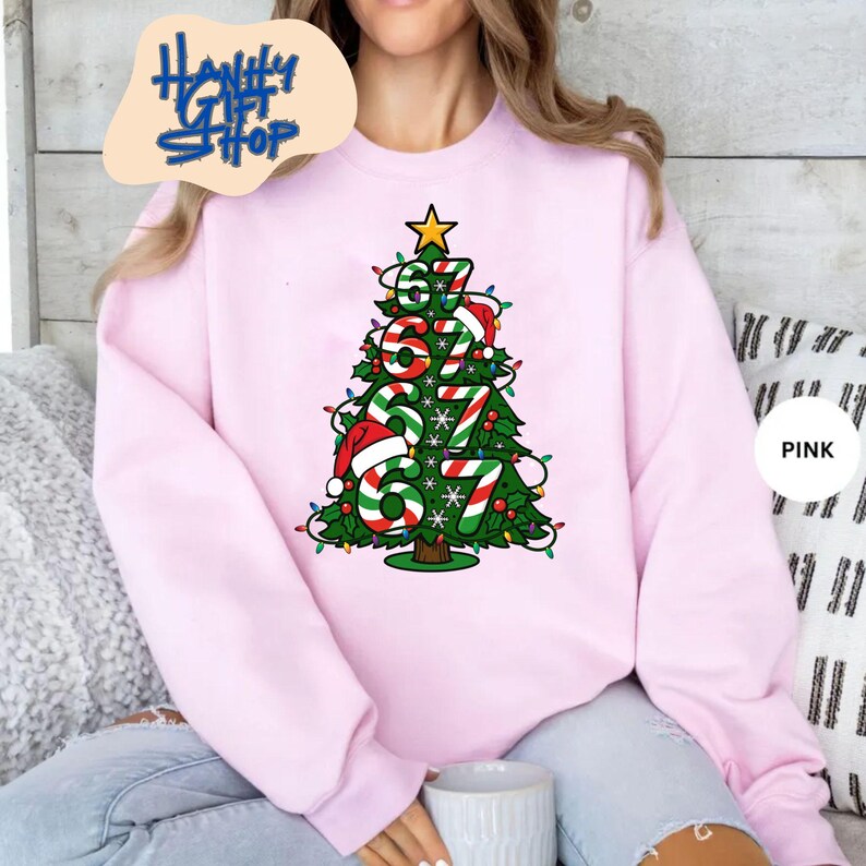 May include: A light pink sweatshirt featuring a Christmas tree design. The tree is composed of the numbers "67" in red, green, and white, adorned with a star and Christmas lights. The sweatshirt has the text "Haniffy Gift Shop" on the top left.