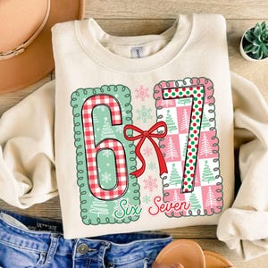 May include: Cream-colored sweatshirt with a festive design featuring the numbers "6" and "7" in a Christmas theme. The numbers are in green and red, with a red bow and the words "Six Seven" below.