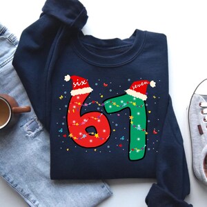 May include: Navy blue sweatshirt with a festive design. The number "61" is displayed in red and green, adorned with Christmas lights and Santa hats. The word "six" and "seven" are written above the numbers.