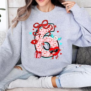 May include: A light gray sweatshirt featuring a large number 6 design. The number is decorated with a pink and white checkered pattern, hearts, a red bow, and a heart-shaped character wearing sunglasses. Additional elements include a gift box, lips, and an envelope with wings.