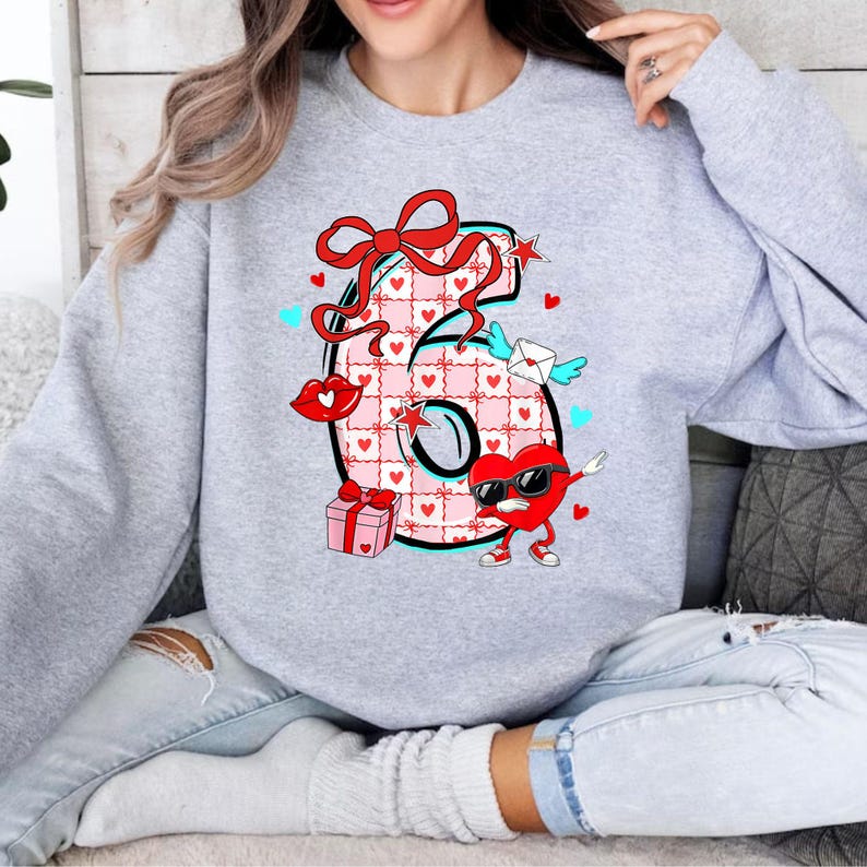 May include: A light gray sweatshirt featuring a large number 6 design. The number is decorated with a pink and white checkered pattern, hearts, a red bow, and a heart-shaped character wearing sunglasses. Additional elements include a gift box, lips, and an envelope with wings.
