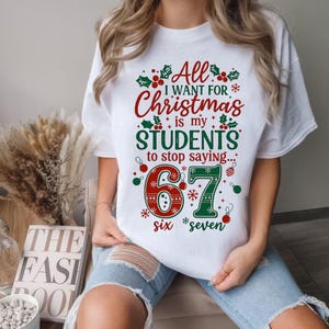 May include: White t-shirt with a Christmas-themed graphic. The design features the text "All I want for Christmas is my students to stop saying..." with the numbers "6" and "7" and the words "six" and "seven". The design includes holly and snowflake accents.