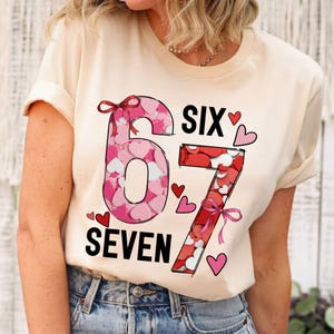 May include: A cream-colored t-shirt featuring the numbers "67" filled with pink and red hearts. The word "SIX" is above the number 6, and "SEVEN" is below the number 7, both in black. Small red hearts are scattered around the numbers.