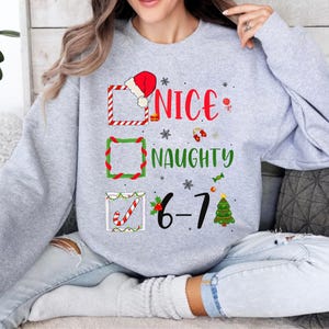 May include: A light gray sweatshirt with a Christmas-themed graphic. The graphic includes the words "NICE" and "NAUGHTY" with check boxes and festive decorations. The number "6-7" is also displayed.