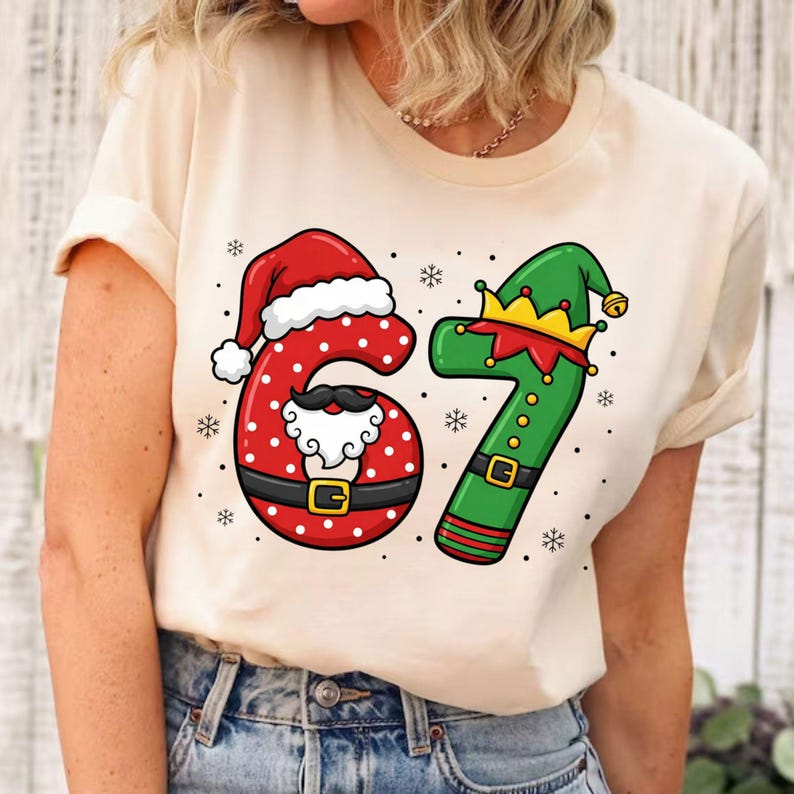 May include: A cream-colored t-shirt featuring the numbers 67 in a festive Christmas design. The number 6 is red with white polka dots, a Santa hat, and a mustache. The number 7 is green with an elf hat and gold accents.
