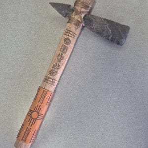 May include: A handcrafted tomahawk with a wooden handle featuring etched designs and a leather grip with a sun symbol. The head is made of dark grey stone, and the blade is a sharp, triangular shape.