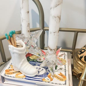 May include: A decorative arrangement featuring white marble-patterned candles, a clear star-shaped candle holder, and a white ceramic boot-shaped match holder filled with matches. The arrangement sits atop a stack of books.