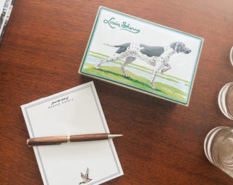 May include: A wooden pen rests on a notepad with the text "from the desk of HUNTER HARRIS" and a duck illustration. A decorative box with a dog illustration and the text "Louis Sherry" is also present.