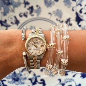 May include: A silver and gold wristwatch with a mother-of-pearl face and diamond hour markers. A silver cuff bracelet and a clear bamboo-style bracelet with gold accents are also visible. The items are displayed on a blue and white patterned surface.