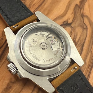May include: Close-up of a stainless steel watch with a transparent back, revealing the intricate mechanical movement. The watch has a black leather strap with tan accents. The names "Adrian", "Sandra", and "Josh" are engraved on the watch face.