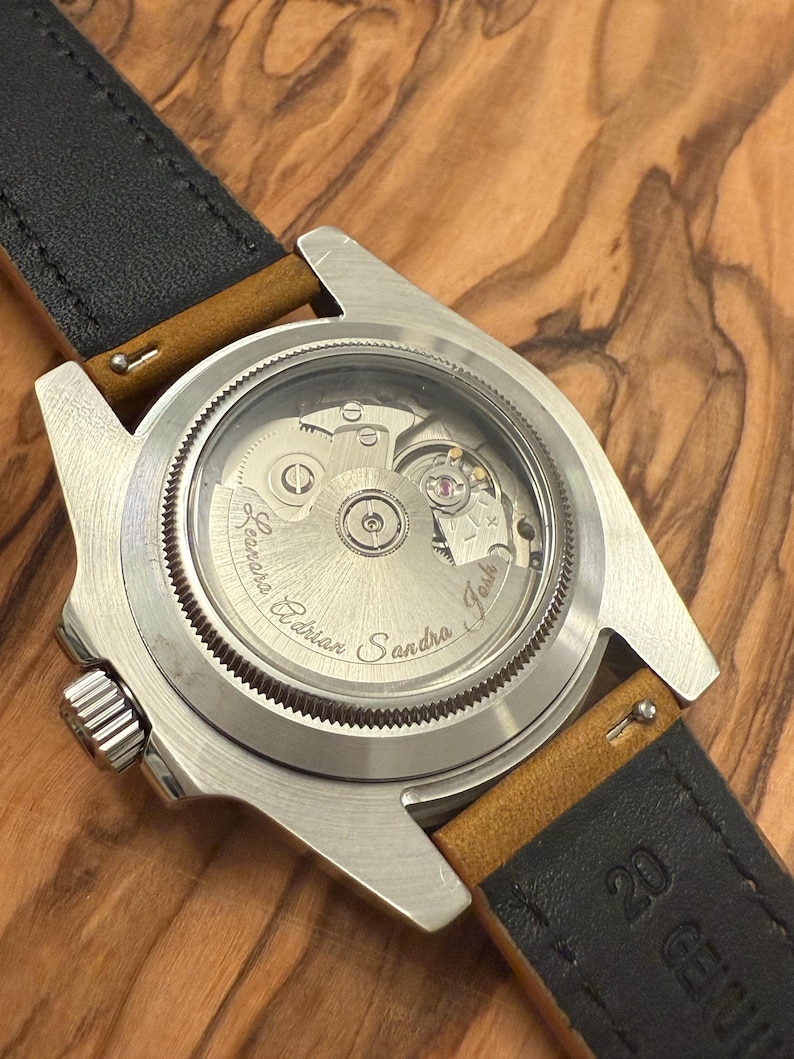 May include: Close-up of a stainless steel watch with a transparent back, revealing the intricate mechanical movement. The watch has a black leather strap with tan accents. The names "Adrian", "Sandra", and "Josh" are engraved on the watch face.