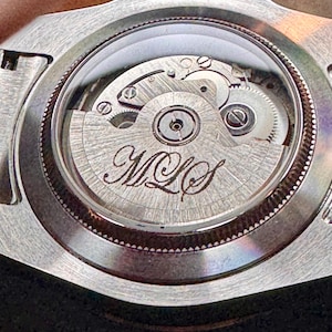 May include: Close-up of the back of a silver wristwatch, showcasing the intricate internal mechanism. The watch's gears and components are visible through a clear glass back. The initials "MLS" are elegantly engraved on the central rotor.