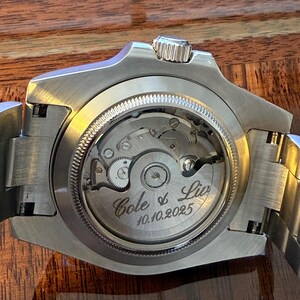 May include: Close-up of a silver wristwatch with a visible mechanical movement. The watch back is engraved with "Cole & Liv" and the date "10.10.2025". The watch has a silver band and a textured crown.
