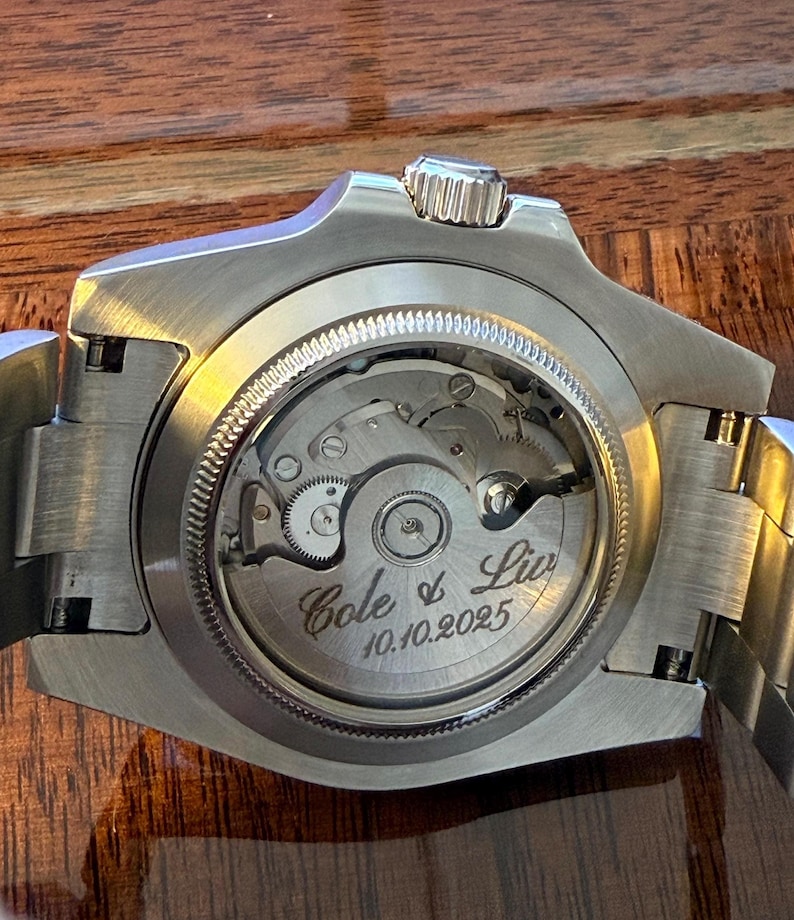 May include: Close-up of a silver wristwatch with a visible mechanical movement. The watch back is engraved with "Cole & Liv" and the date "10.10.2025". The watch has a silver band and a textured crown.