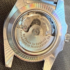May include: Close-up of a silver-toned watch back, showcasing the intricate internal mechanism. The watch features the text "80 Years in Service Middleburg, VA" around the edge. The watch's gears and components are visible through the clear back.