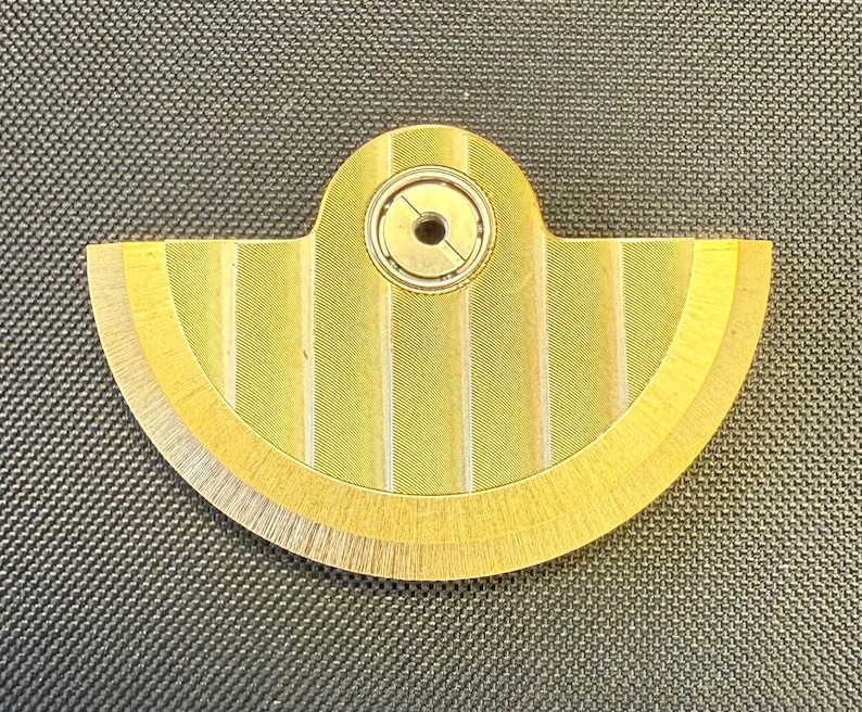 May include: A gold-colored, semi-circular mechanical component with a central circular bearing. The component has a fan-like design with vertical striped detailing and a textured surface. The overall shape is reminiscent of a stylized fan or gear.