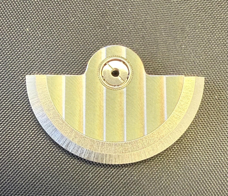May include: A fan-shaped, metallic object with a central circular component and a series of vertical stripes. The object has a gold and silver color scheme, with a brushed metal finish on the edges. The design is symmetrical and appears to be a mechanical part.