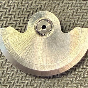 May include: A close-up of a silver-colored watch part with a curved, fan-like shape. The part has a central circular component with a small hole and intricate inner workings. The surface has a brushed texture, and the edges are smooth and rounded.