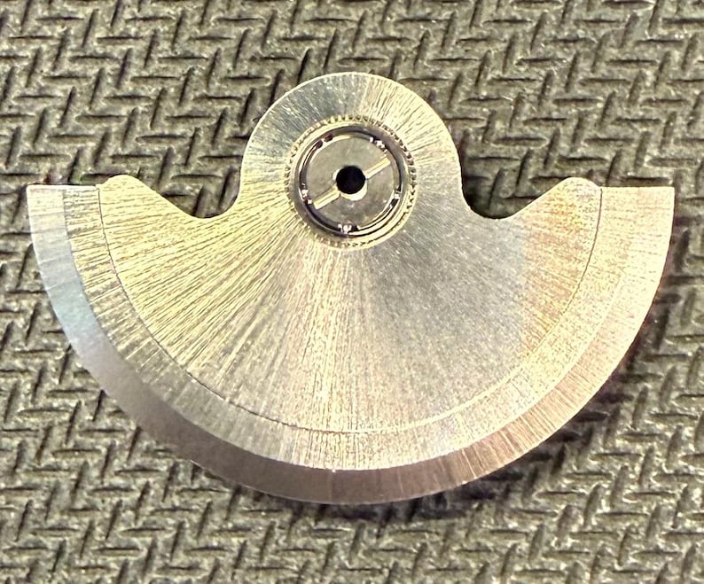 May include: A close-up of a silver-colored watch part with a curved, fan-like shape. The part has a central circular component with a small hole and intricate inner workings. The surface has a brushed texture, and the edges are smooth and rounded.