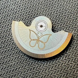 May include: A metal watch part with a butterfly design. The part is silver with gold accents and a circular element at the top. The butterfly is etched in gold glitter.