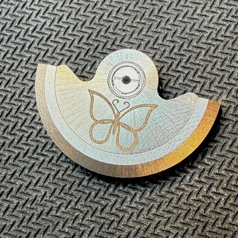May include: A metal watch part with a butterfly design. The part is silver with gold accents and a circular element at the top. The butterfly is etched in gold glitter.