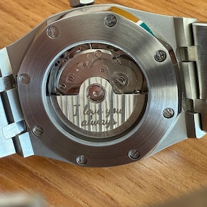 May include: Close-up of a silver-toned watch with a visible mechanical movement. The watch has a round window on the back, revealing intricate gears and the engraved phrase "I love you always". The watch is on a wooden surface.