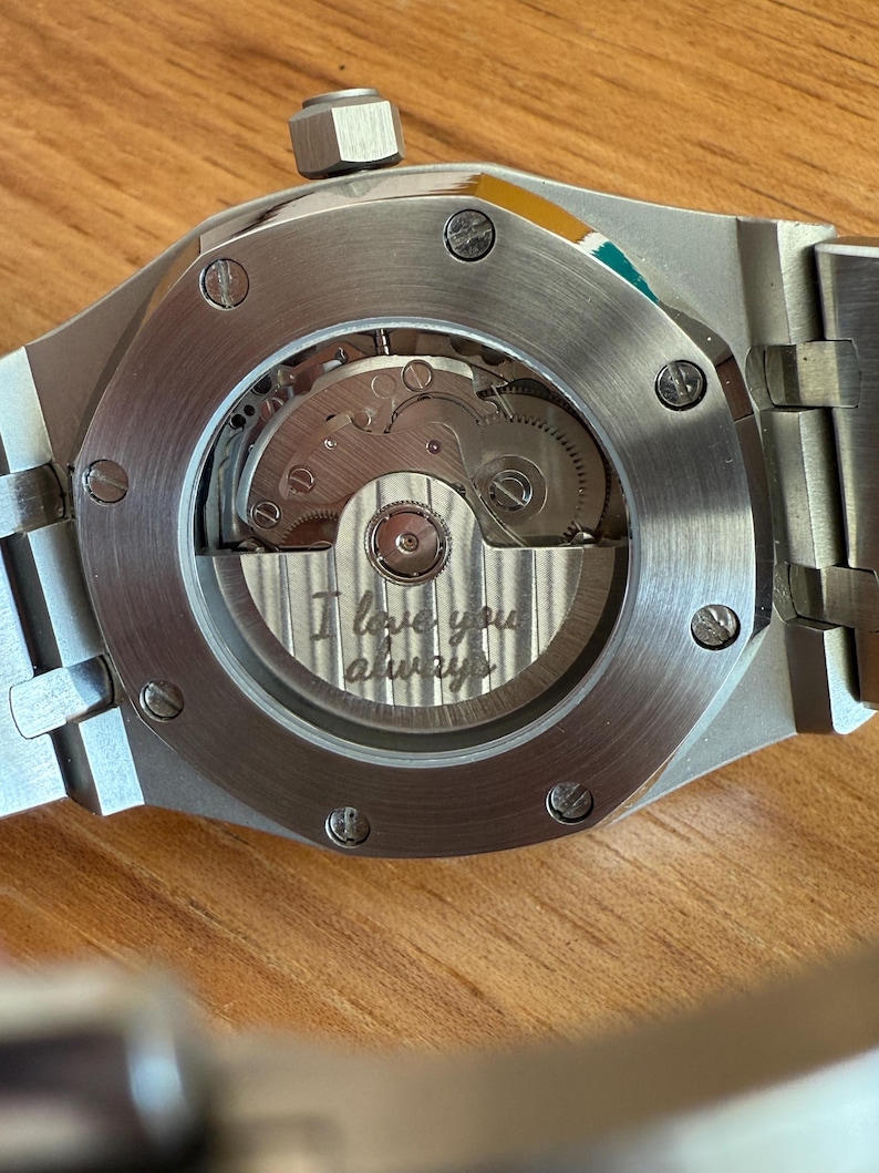 May include: Close-up of a silver-toned watch with a visible mechanical movement. The watch has a round window on the back, revealing intricate gears and the engraved phrase "I love you always". The watch is on a wooden surface.