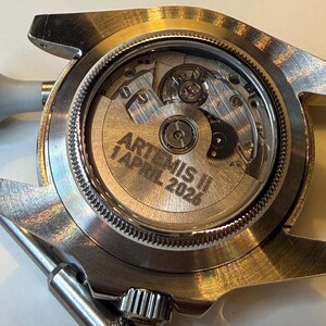 May include: Close-up of a silver-toned watch with a transparent back, revealing the intricate mechanical movement. The watch face displays the text "ARTEMIS II 1 APRIL 2026". The watch has a silver bezel and a metal band.