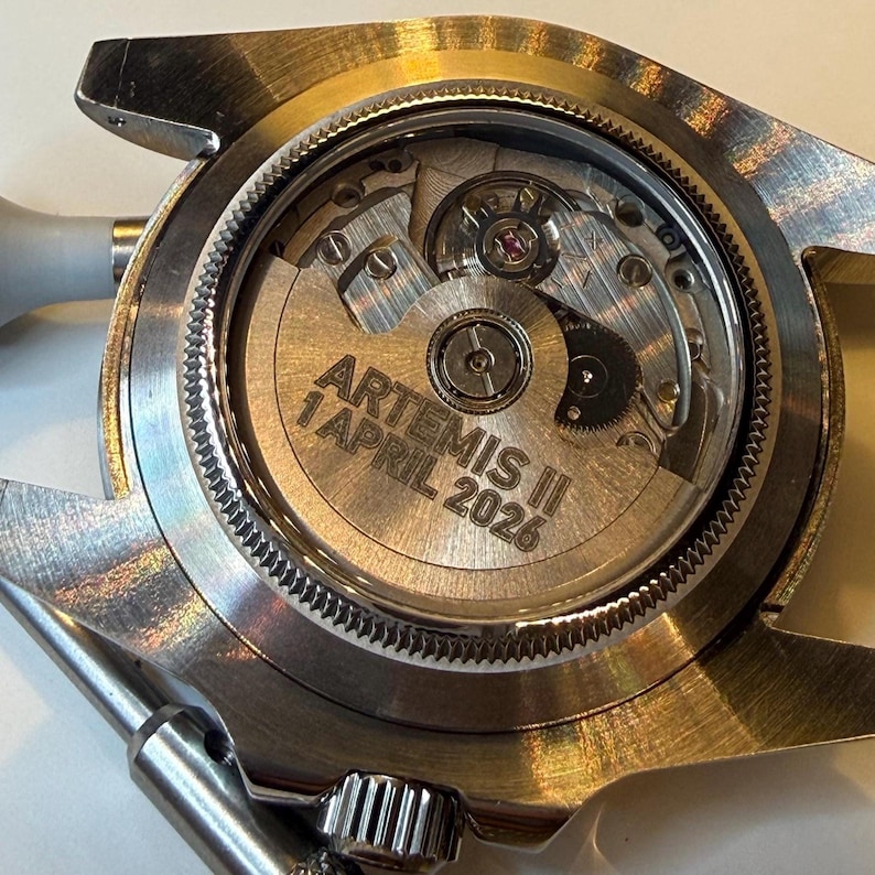 May include: Close-up of a silver-toned watch with a transparent back, revealing the intricate mechanical movement. The watch face displays the text "ARTEMIS II 1 APRIL 2026". The watch has a silver bezel and a metal band.