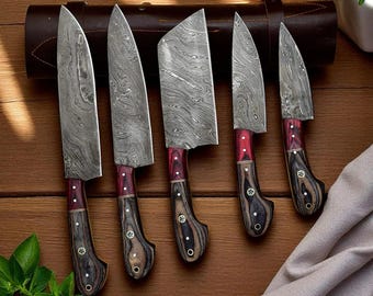 Puede incluir: Un juego de cinco cuchillos de cocina de acero de Damasco con hojas estampadas y mangos rojos y de madera. Los cuchillos están colocados sobre una superficie de madera, con un rollo de cuchillos de cuero en el fondo.