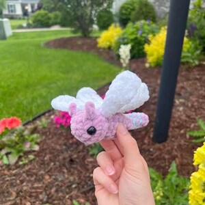 May include: A handmade crochet butterfly in shades of pink, white, and blue. The butterfly has a fluffy texture, with white wings and a pink body. The wings are large and detailed, and the body has a small black eye. The butterfly is held in a hand.
