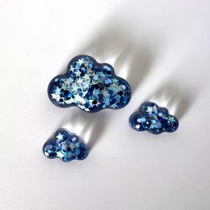 May include: Three cloud-shaped resin pieces filled with blue and silver glitter and star-shaped confetti. The clouds vary in size, with a dark blue outline. The pieces are arranged on a white surface.
