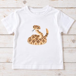 May include: A white short-sleeved t-shirt featuring a centered watercolor-style illustration of a coiled rattlesnake. The snake is depicted in shades of tan and brown with a distinct diamond pattern on its body and a dark, forked tongue extended.