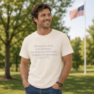 May include: Cream-coloured t-shirt with the text "We hold these truths to be self-evident, that all men are created equal... - Declaration of Independence, July 4, 1776." The shirt is worn outdoors with an American flag in the background.
