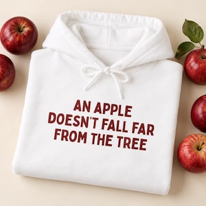 Puede incluir: Una sudadera con capucha blanca que presenta el texto rojo oscuro AN APPLE DOESN'T FALL FAR FROM THE TREE impreso en el frente. La sudadera tiene una capucha con cordón y está rodeada de varias manzanas rojas enteras con hojas verdes.
