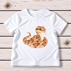 Könnte beinhalten: Ein weißes T-Shirt mit kurzen Ärmeln und einer mittigen Aquarell-Illustration einer zusammengerollten braun-beigen Schlange, deren kleine rosa Zunge aus dem Mund ragt. Die Schlange hat große dunkle Augen und einen gemusterten, segmentierten Körper.
