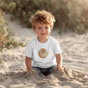 May include: A white short-sleeved t-shirt featuring a centered graphic of a brown and tan nautilus shell. The shell design displays a detailed spiral pattern with natural earth tones, printed on the front of the plain white cotton fabric.