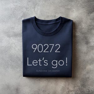 May include: Navy blue crew neck t-shirt with the text "90272 Let's go!" in white, along with "SUNSHINE ON WATER" below. The shirt is folded neatly on a gray surface.