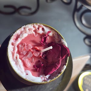 Pode incluir: Uma vela pequena e redonda numa lata dourada. A vela tem um centro de cera rosa, bordas brancas e é decorada com cristais transparentes e pétalas de rosa secas. Um pavio branco é visível.