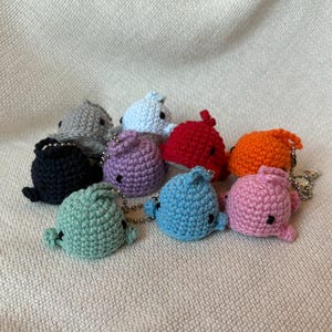 Könnte beinhalten: Eine Sammlung handgefertigter Wal-Schlüsselanhänger aus Häkelarbeit in verschiedenen Farben, darunter Grau, Blau, Rot, Orange, Lila, Grün und Rosa. Jeder Wal hat kleine schwarze Augen und eine silberne Kette.