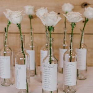 May include: Clear glass bottles with white labels and white roses. Each bottle holds a single white rose, creating a simple floral arrangement. The labels have names, ideal for a wedding or event.