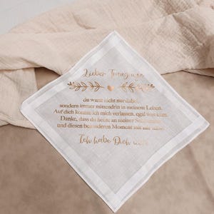 Könnte beinhalten: Weißes Taschentuch mit goldfarbener Inschrift und dekorativen floralen Elementen. Der Text ist auf Deutsch. Das Taschentuch liegt auf einem beigen Stoff.