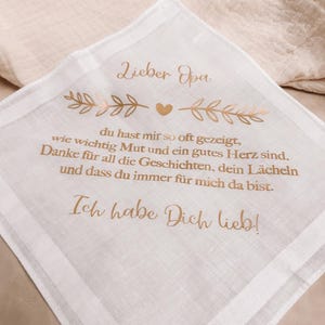Könnte beinhalten: Weißes Taschentuch mit goldenem Text und dekorativen Blatt- und Herzakzenten. Der Text lautet "Lieber Opa" und enthält eine liebevolle Botschaft. Ein durchdachtes Geschenk für einen Großelternteil.