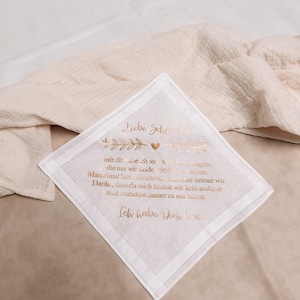 Könnte beinhalten: Ein weißes Taschentuch mit goldenem Text und einem dekorativen Rand. Der Text enthält den Satz "Liebe Schwester" und andere deutsche Texte. Das Taschentuch liegt auf einer beigen Oberfläche.