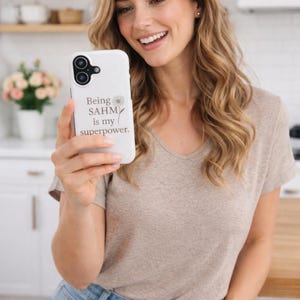 Peut inclure: Coque de téléphone blanche avec le texte "Being SAHM is my superpower" et un motif de marguerite. La coque est tenue par une personne portant un t-shirt beige à col en V et un jean bleu.