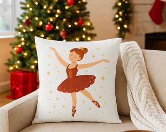 May include: A white square throw pillow featuring a cartoon ballerina in a burgundy dress and ballet shoes. The ballerina has a gold crown and is surrounded by gold stars. The pillow is on a beige couch in front of a Christmas tree.